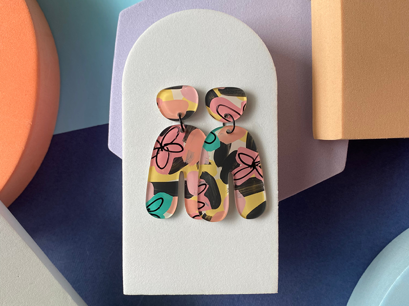 A pair of abstract earrings displayed on a white arch-shaped stand. The earrings are made of a glossy material and feature a bold, colourful pattern with pink, yellow, black, teal, and coral brushstrokes and line art. The background includes soft geometric shapes in pastel tones of lavender, peach, and navy.
