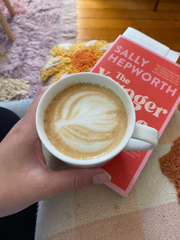 Hand holding a cup of coffee with latte art, resting on a book titled “The Younger Wife” by Sally Hepworth, cosy setting with textured rug and blanket.
