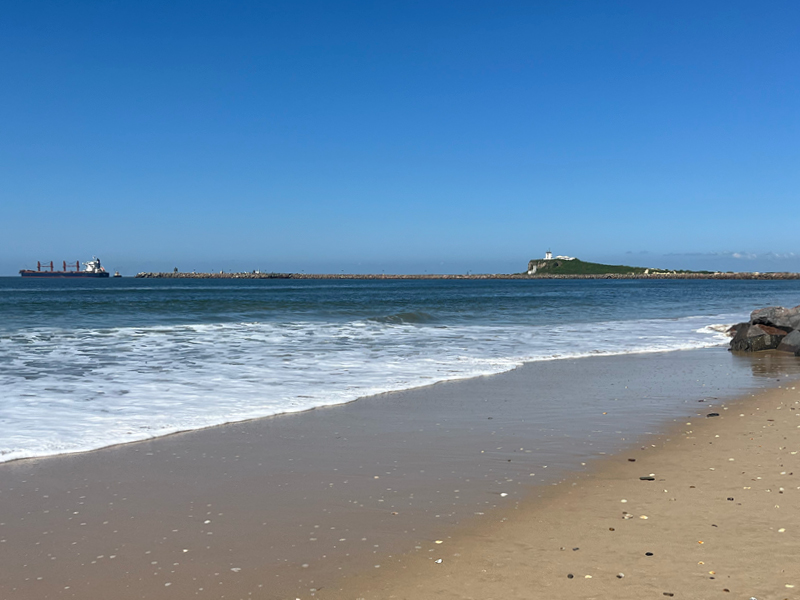 Slow living moment on Stockton beach with ocean waves, Nobby’s Head lighthouse and a cargo ship on the horizon of Newcastle.