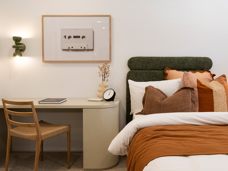 Modern bedroom styled by Mojo Homes with green bedhead, warm-toned cushions, and a minimalist desk for a cosy, functional design.