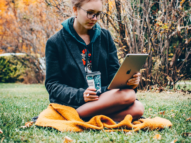 A woman sitting on a blanket outdoors in autumn, reading on a tablet and holding a coffee cup – a relaxed scene symbolising slow tech living.