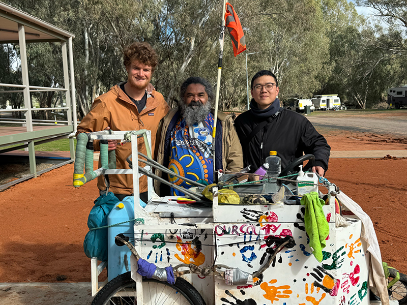 A group of three people standing beside a hand-painted cart covered in colourful handprints in an outback setting, featured as part of a story connected to the 2025 Newcastle Citizen of the Year.