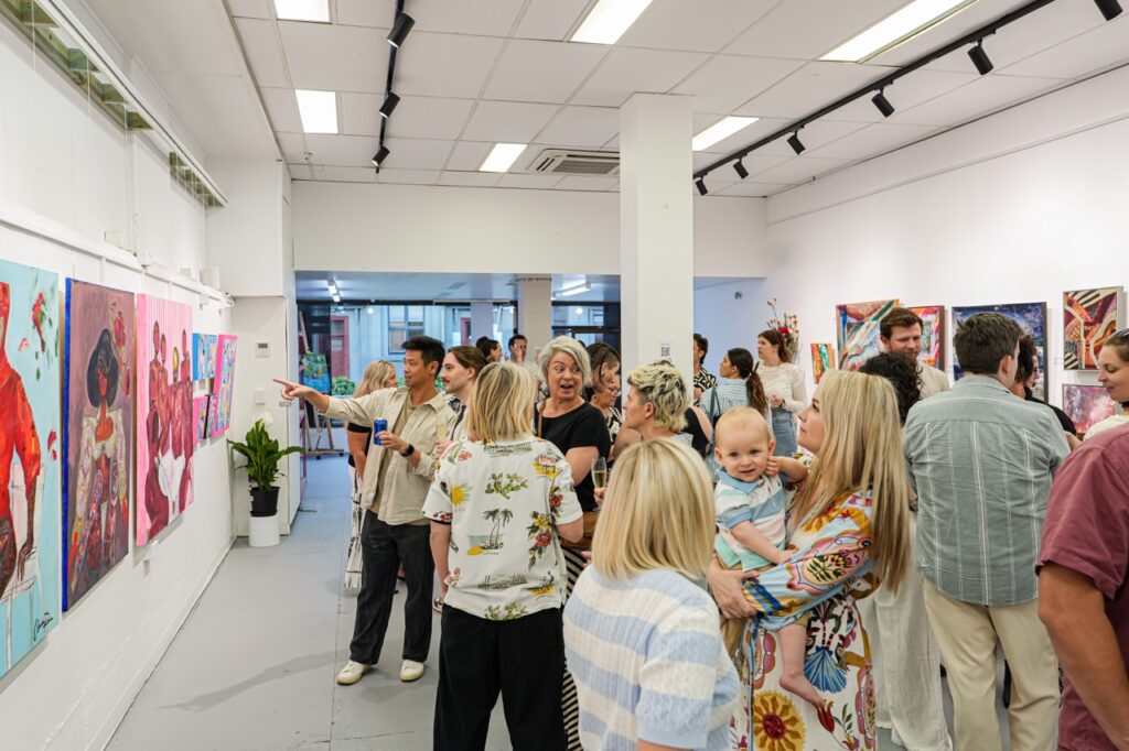 A large crowd gathers inside Alejandro’s gallery during an exhibition opening, with people viewing colourful artworks displayed along the walls.