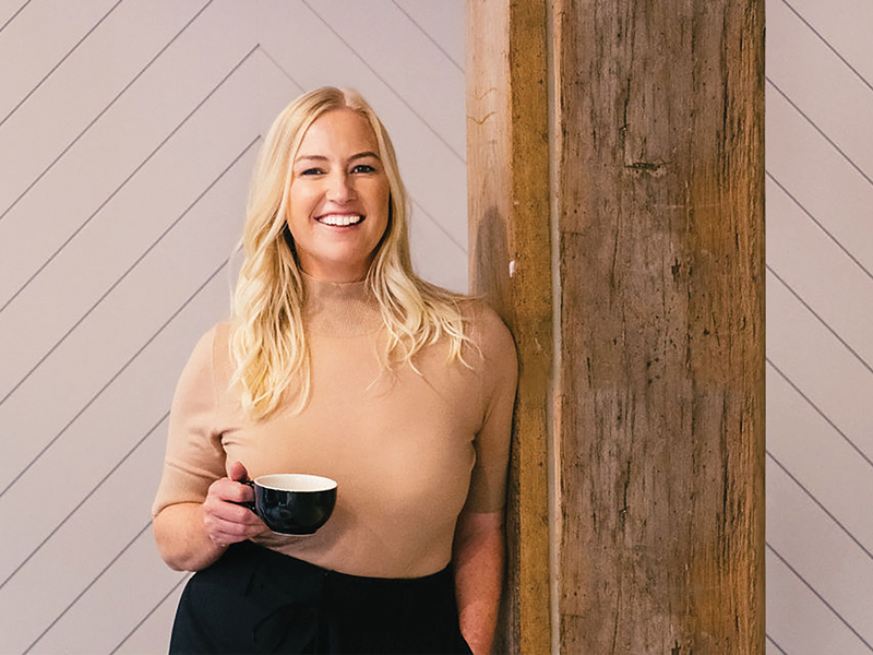 A woman holding a coffee cup and smiling while standing indoors beside a timber post.