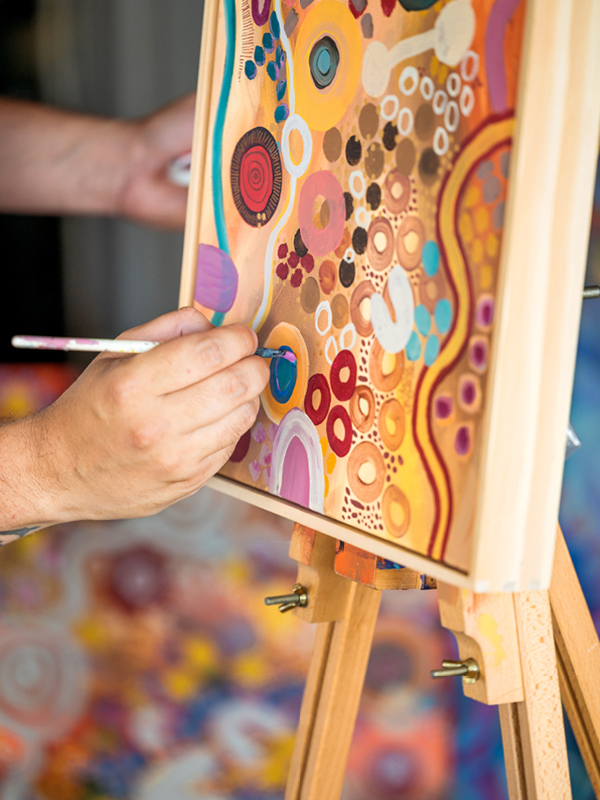 A close-up of an artist’s hand painting a vibrant artwork filled with colourful circular patterns and organic shapes on a canvas positioned on an easel.