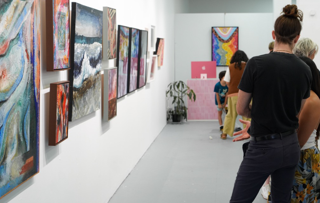 A row of colourful artworks displayed along a white gallery wall at Alejandro’s, with visitors viewing the exhibition in the background.