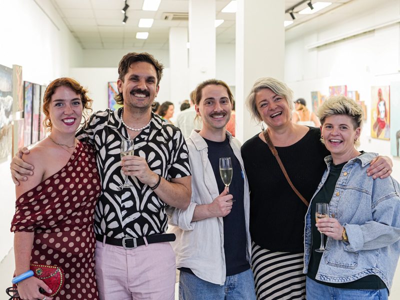 A group of people stand together smiling and holding drinks inside Alejandro’s gallery during an exhibition opening, with colourful artworks displayed on the walls behind them.